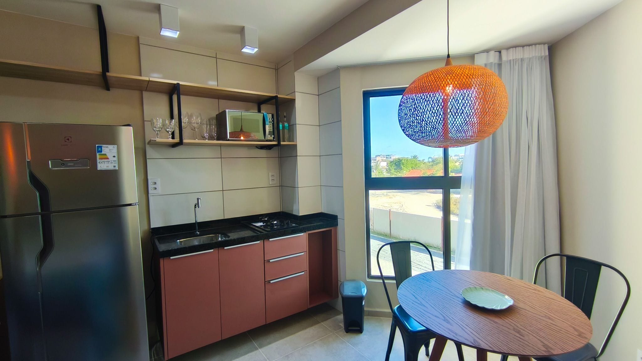 A modern kitchen with a refrigerator, stove, and sink, a dining table with chairs, and a large window with a view.