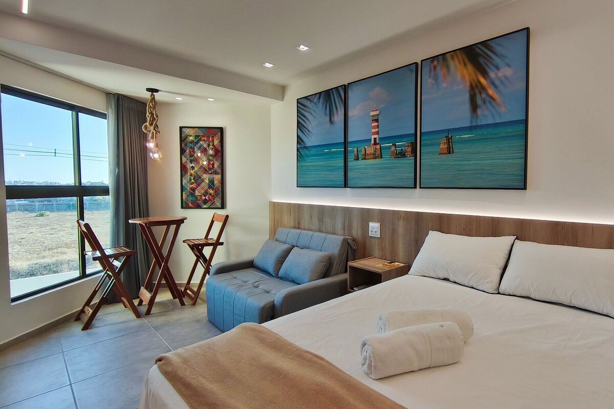 A hotel room with a bed, a seating area with chairs, a small table, a painting of a beach scene, and a view of the outdoors.