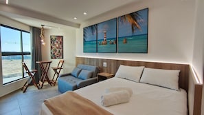 A hotel room with a bed, a seating area with chairs, a small table, a painting of a beach scene, and a view of the outdoors.