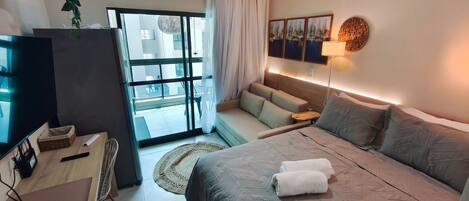 A modern hotel room with a bed, a TV, a desk, and a view of the outside.