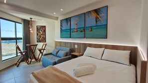 A hotel room with a bed, a seating area with chairs, a small table, a painting of a beach scene, and a view of the outdoors. - Max Carneiros Suites by CTA (Tamandaré)