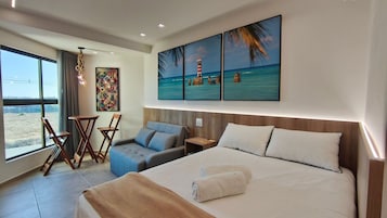 Quarto de hotel com uma cama, área de estar com cadeiras, uma mesinha e uma pintura de uma paisagem de praia, além de vista para o exterior.