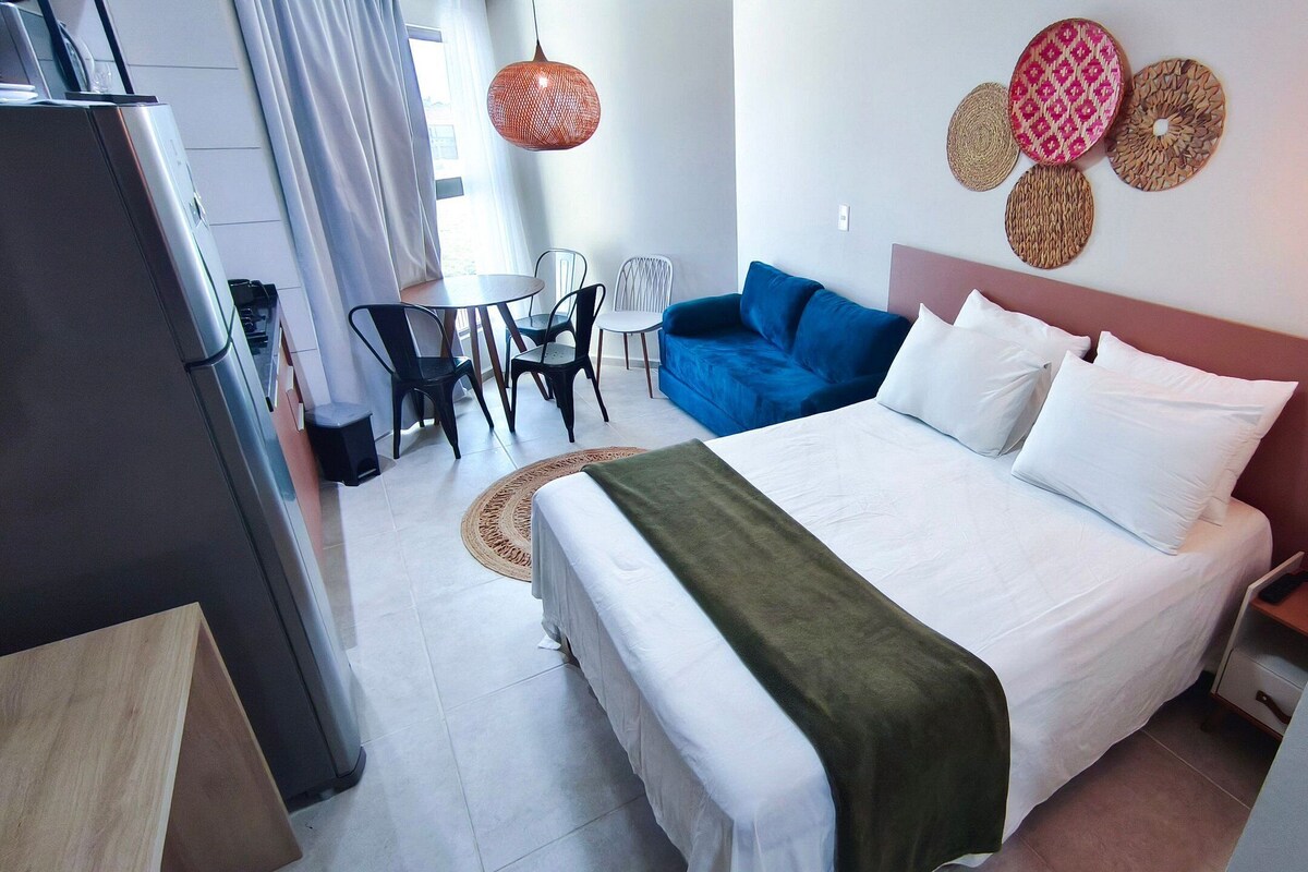 A hotel room with a bed, a blue sofa, a dining table with chairs, a small kitchen area, and decorative wall art.
