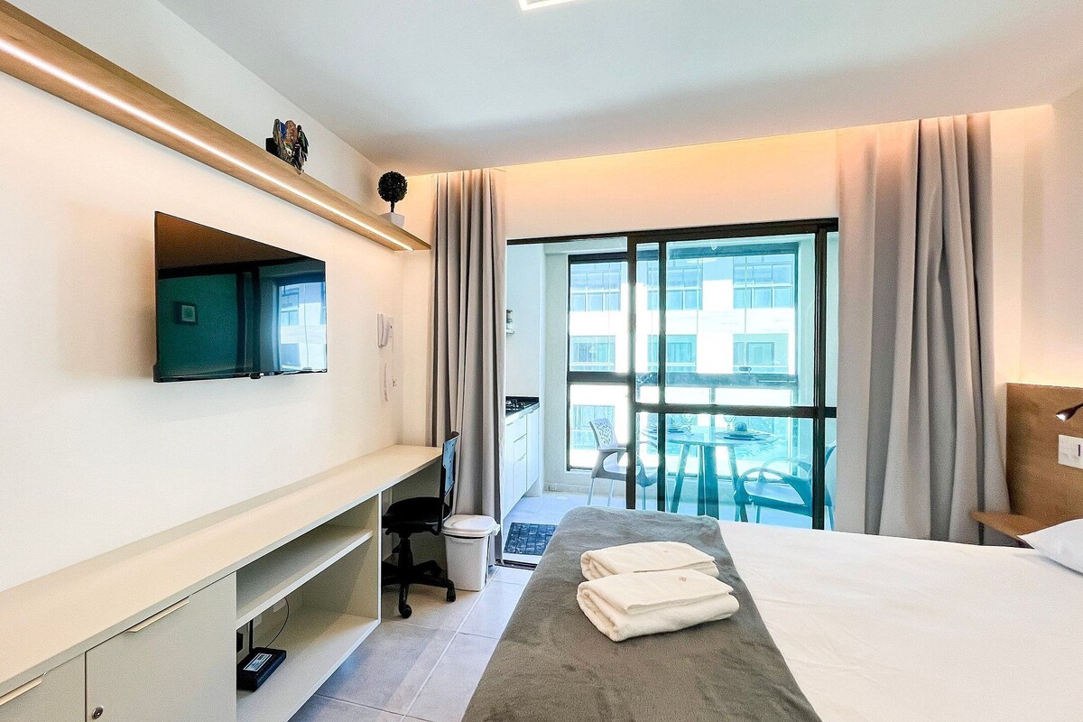 A hotel room with a bed, a desk, a chair, a TV, a window with curtains, and a balcony with outdoor furniture.