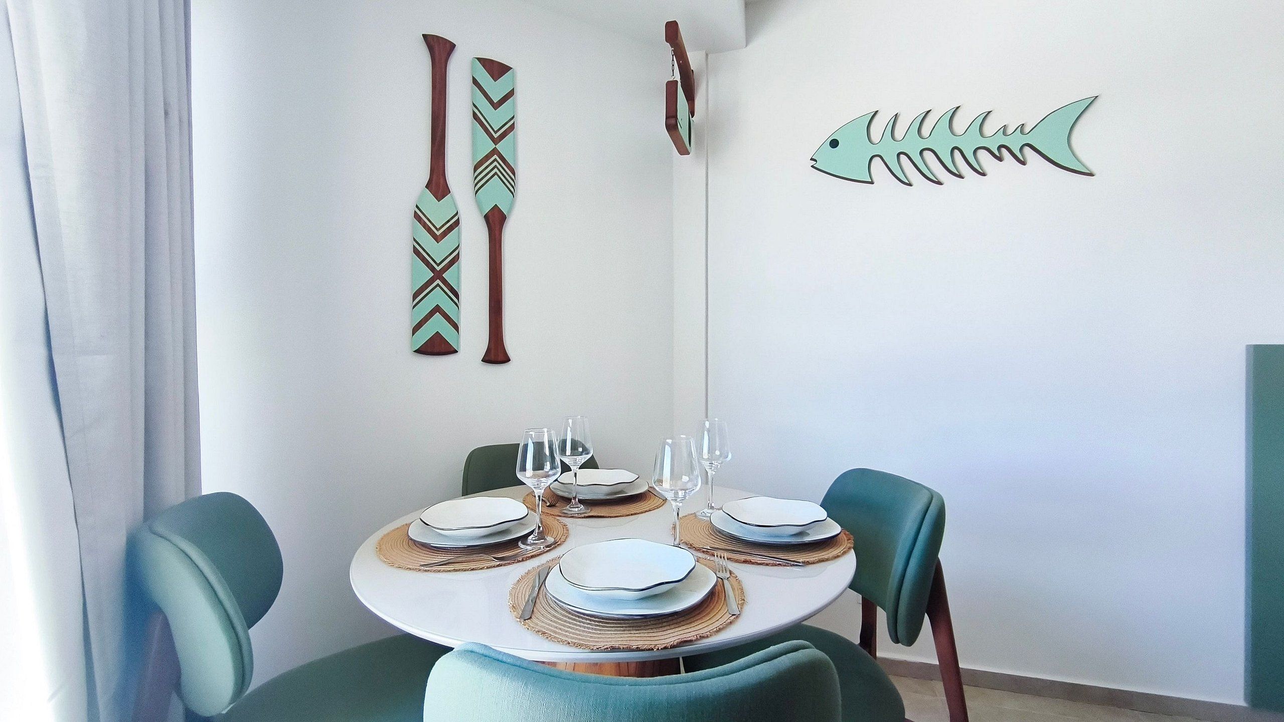 Una zona de menjador amb una taula rodona preparada per a quatre, amb cadires color turquesa, una decoració de peix penjada a la paret i dos penjals.