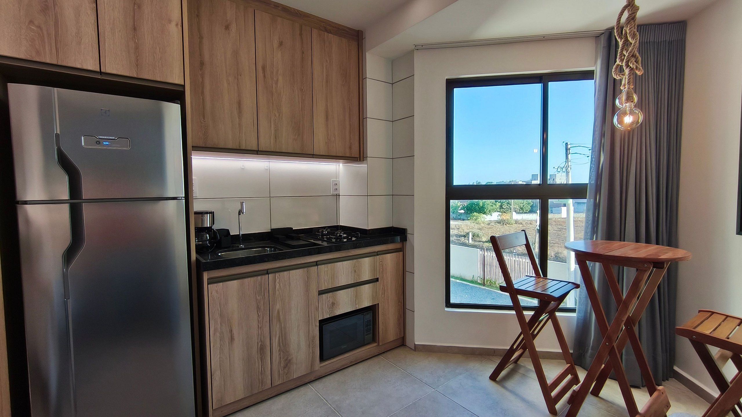 Een moderne keuken met houten kastjes, een roestvrijstalen koelkast, een gasfornuis en een magnetron. Er is een eethoek met een houten tafel en stoelen, en een groot raam met uitzicht naar buiten.