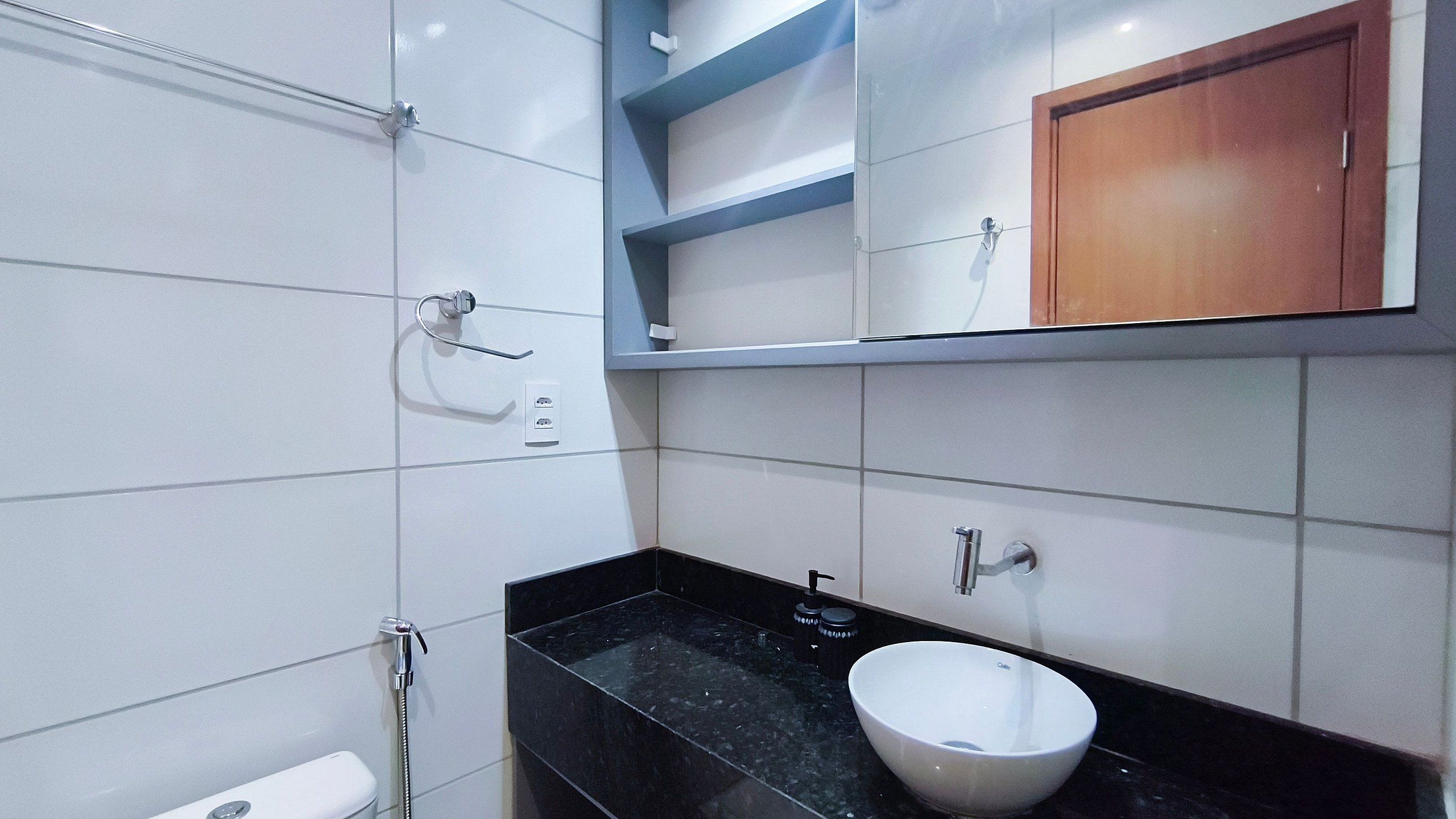 Ein modernes Badezimmer mit weißen Fliesen, einer schwarzen Arbeitsplatte, einem weißen Waschbecken und einem Holzkabinett über dem Spiegel.