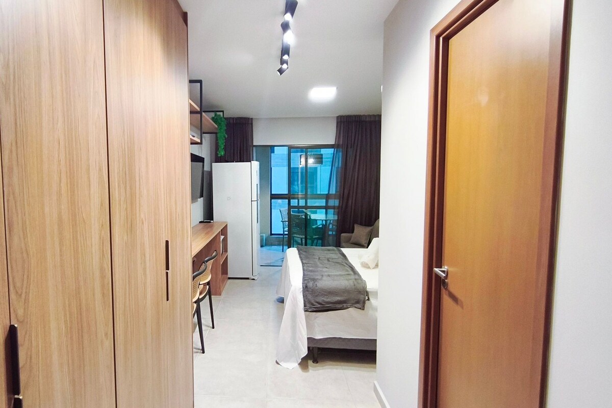 A narrow hallway leading to a bedroom with a bed, a desk, and a refrigerator.