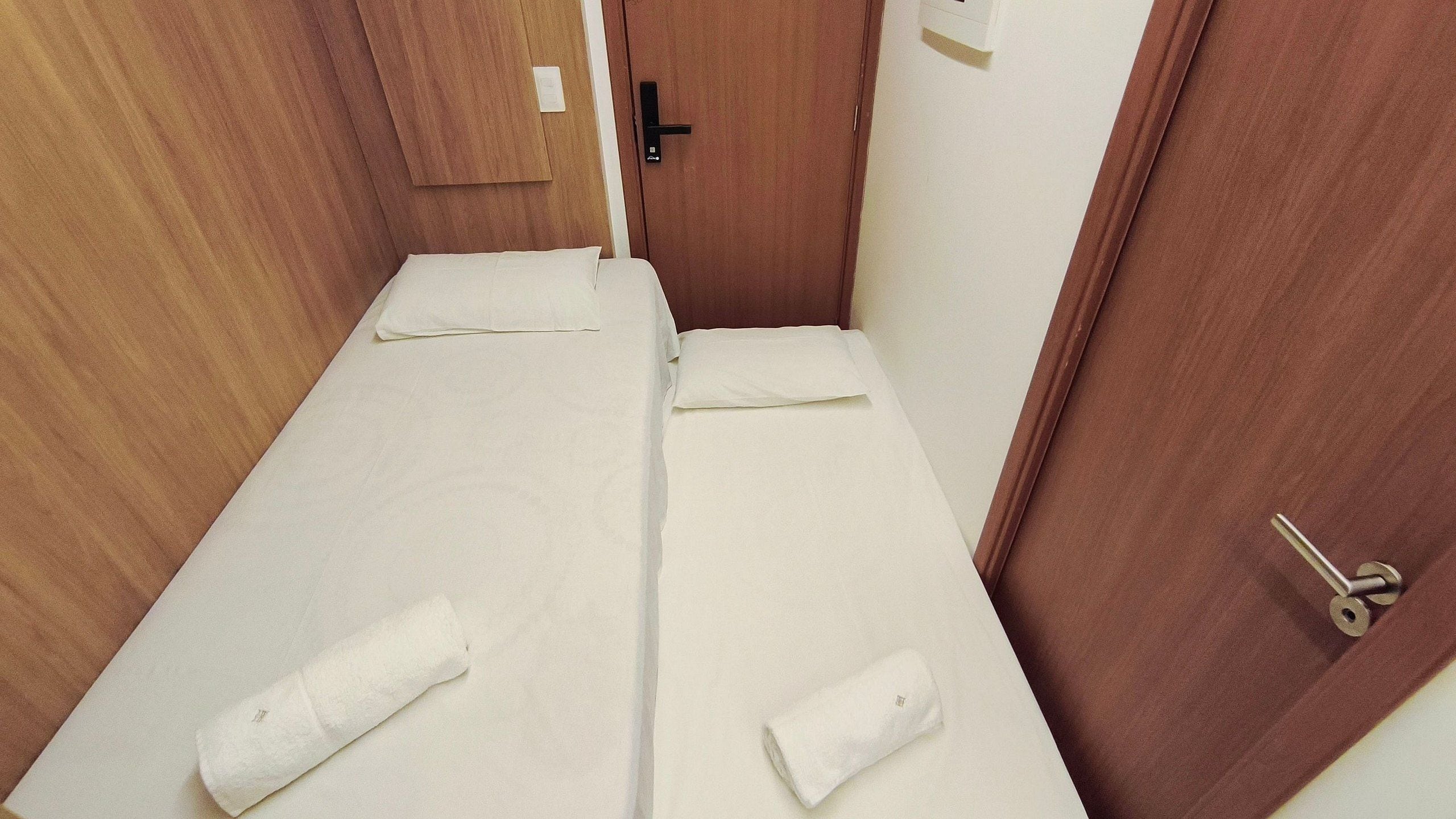 Ein kleiner, schmaler Raum mit einer Holztür, einem Bett mit weißer Bettwäsche und einem Handtuch auf dem Bett.