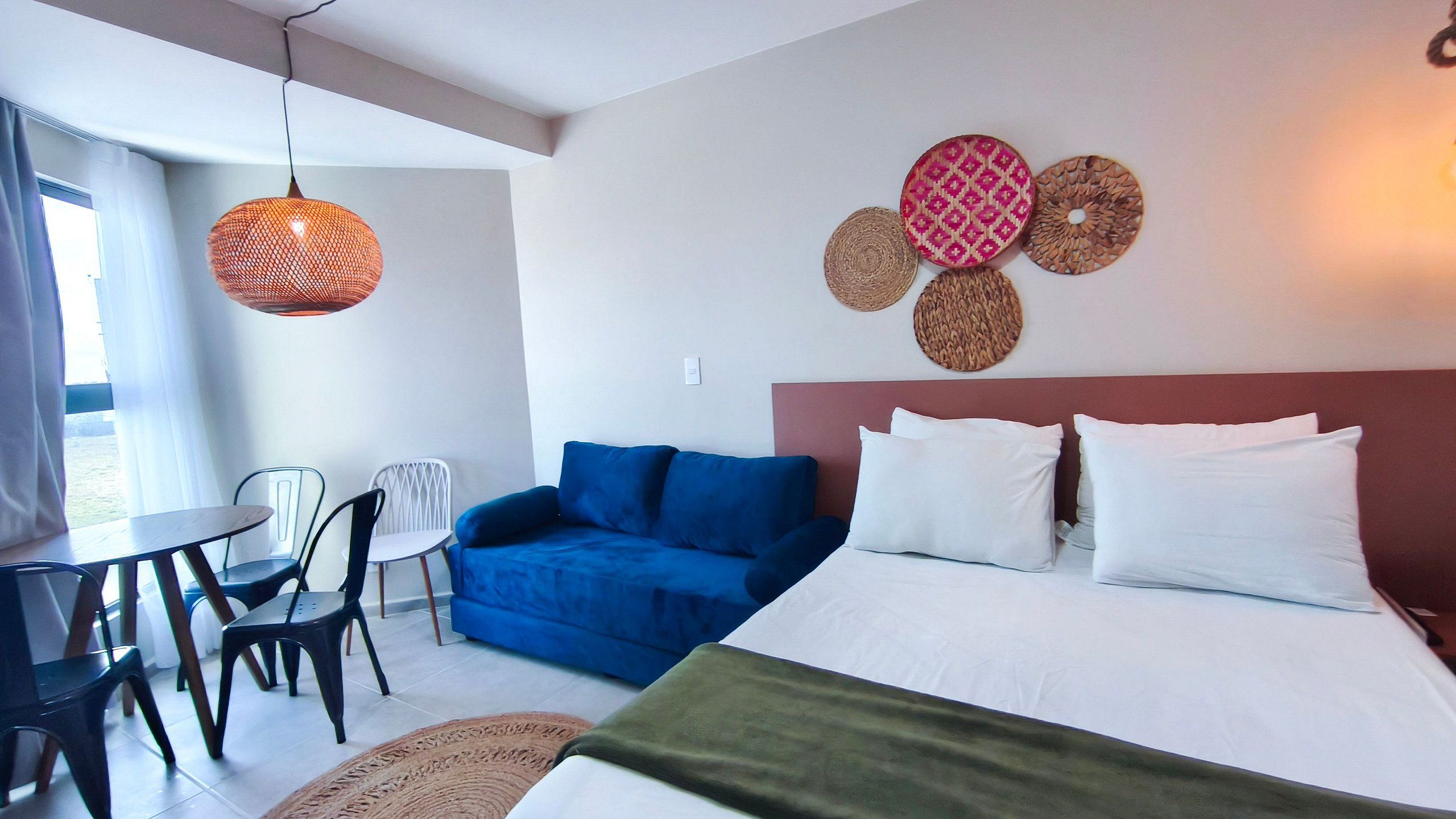 A hotel room with a bed, blue sofa, white pillows, a dining table with chairs, and a hanging light fixture.