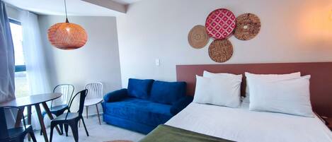 A hotel room with a bed, blue sofa, white pillows, a dining table with chairs, and a hanging light fixture.