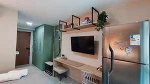 Een compacte kamer met een flatscreen televisie aan de muur, een inbouwkoelkast, een klein zitgedeelte met een bankje, en een deur naar een andere kamer.