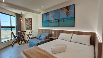 Habitación de hotel con cama, zona de estar con sillas, una mesita y un cuadro de una playa, además de vistas al exterior.