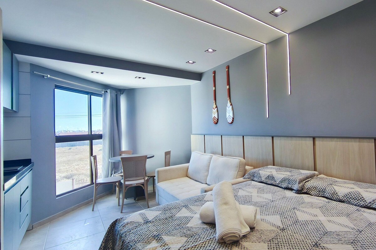 Una habitación de hotel moderna con una cama grande, una zona de estar con una mesa y sillas, vistas a través de un ventanal amplio y una decoración de murciélago en la pared.