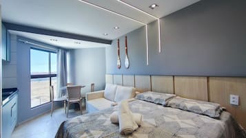 A modern hotel room with a large bed, a seating area with a table and chairs, a view through a large window, and a wall-mounted bat decoration.