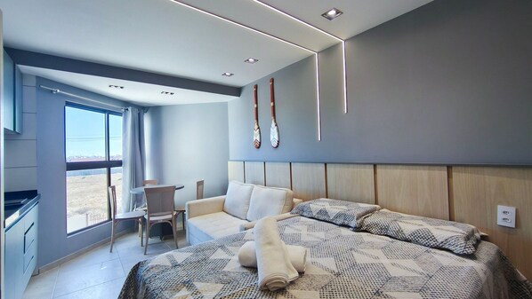 A modern hotel room with a large bed, a seating area with a table and chairs, a view through a large window, and a wall-mounted bat decoration.