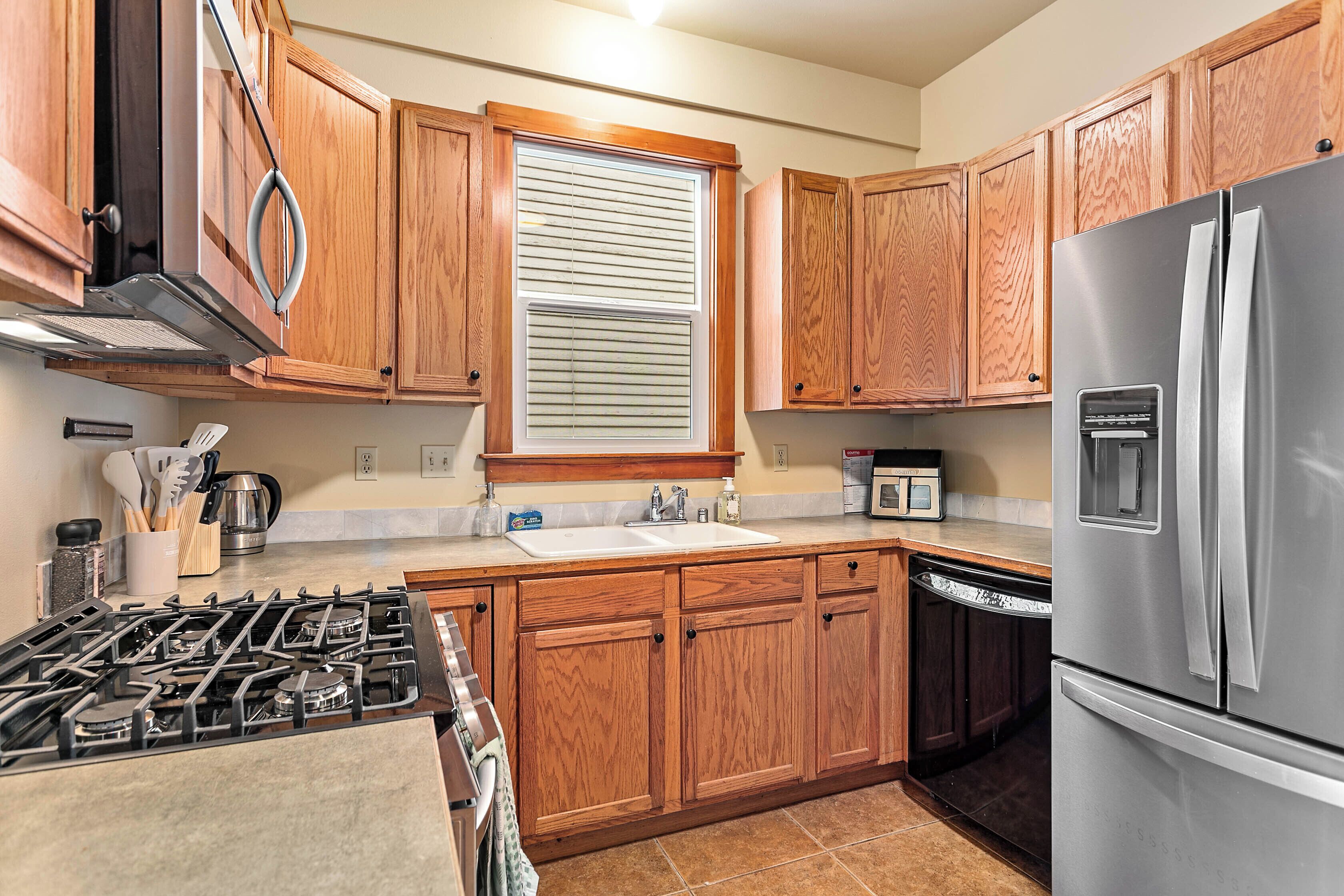 A kitchen with wooden cabinets, a stainless steel refrigerator, a gas stove, a window with blinds, and a microwave.