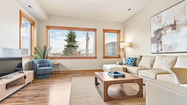 A modern living room with a wooden coffee table, a beige sofa, and a blue armchair. There are large windows with blinds, a view of a tree outside, and a painting on the wall.