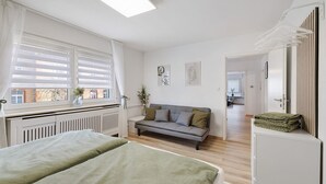 Un dormitorio con cama, sofá, radiador, ventana con persianas y puerta que da a un pasillo.