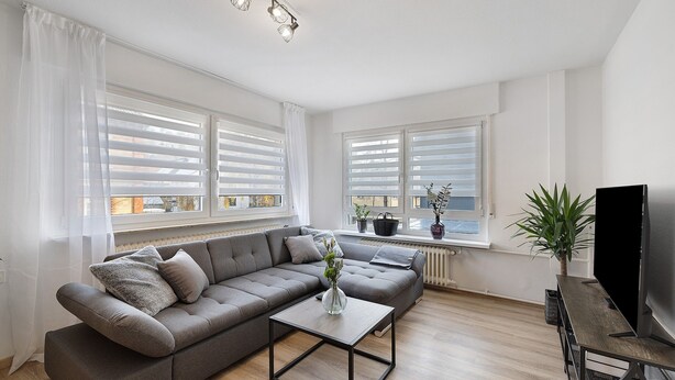Una sala de estar moderna con un sofá seccional gris, una mesa de centro, un televisor de pantalla plana y ventanas grandes con persianas blancas.