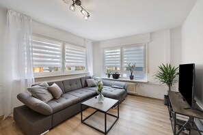Un salon moderne avec un canapé d’angle gris, une table basse, un téléviseur à écran plat et de grandes fenêtres équipées de stores blancs.