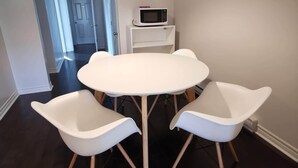 A dining area with a white round table and two white chairs with wooden legs. A white microwave is on a small white shelf against the wall. - Montreal Mile-Ex close to MIL (Montréal)