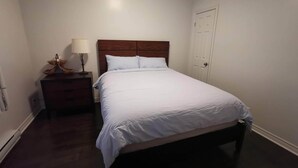 A bedroom with a wooden headboard, a white bedspread, a nightstand with a lamp, and a door. - Montreal Mile-Ex close to MIL (Montréal)