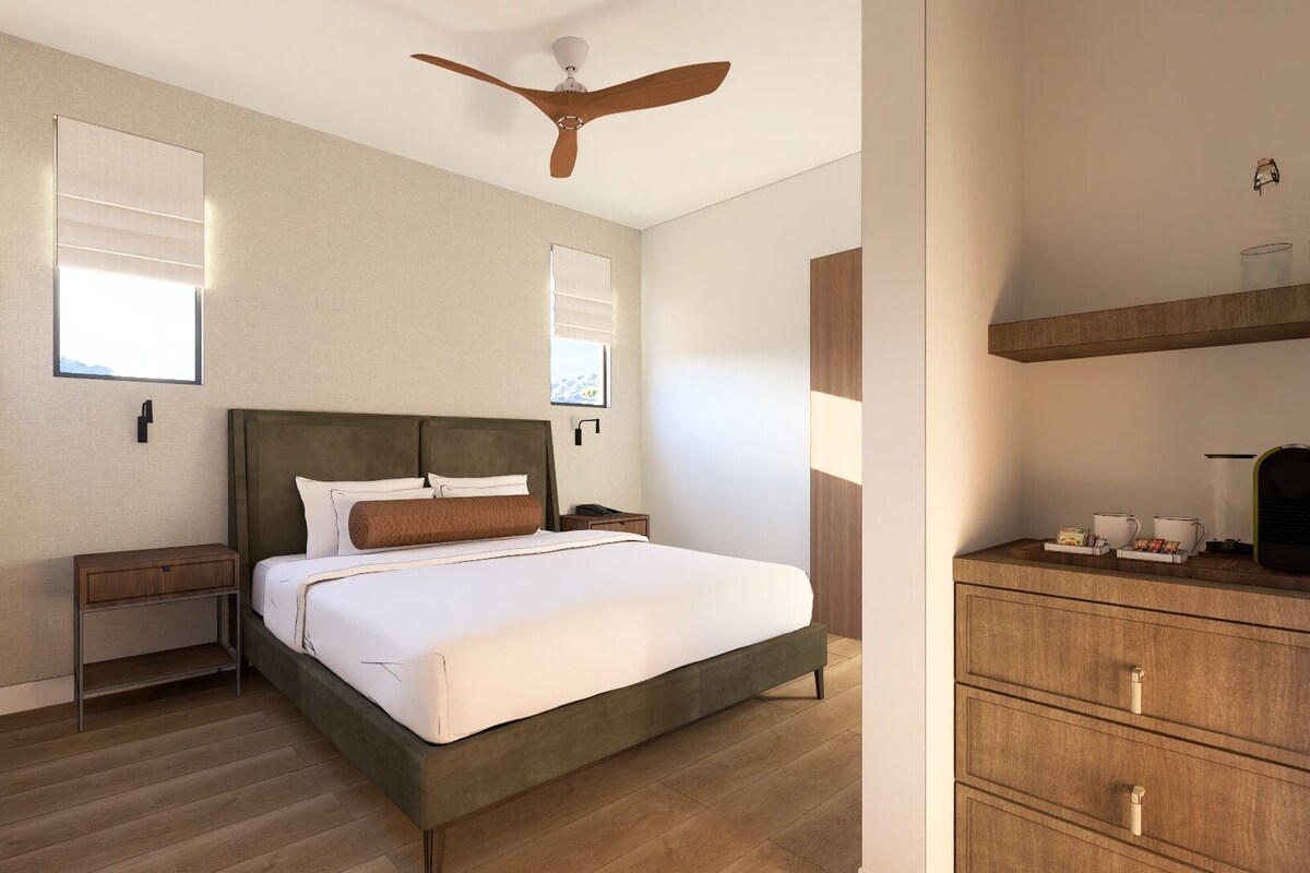 A modern hotel room with a bed, wooden dresser, ceiling fan, and a window with blinds.