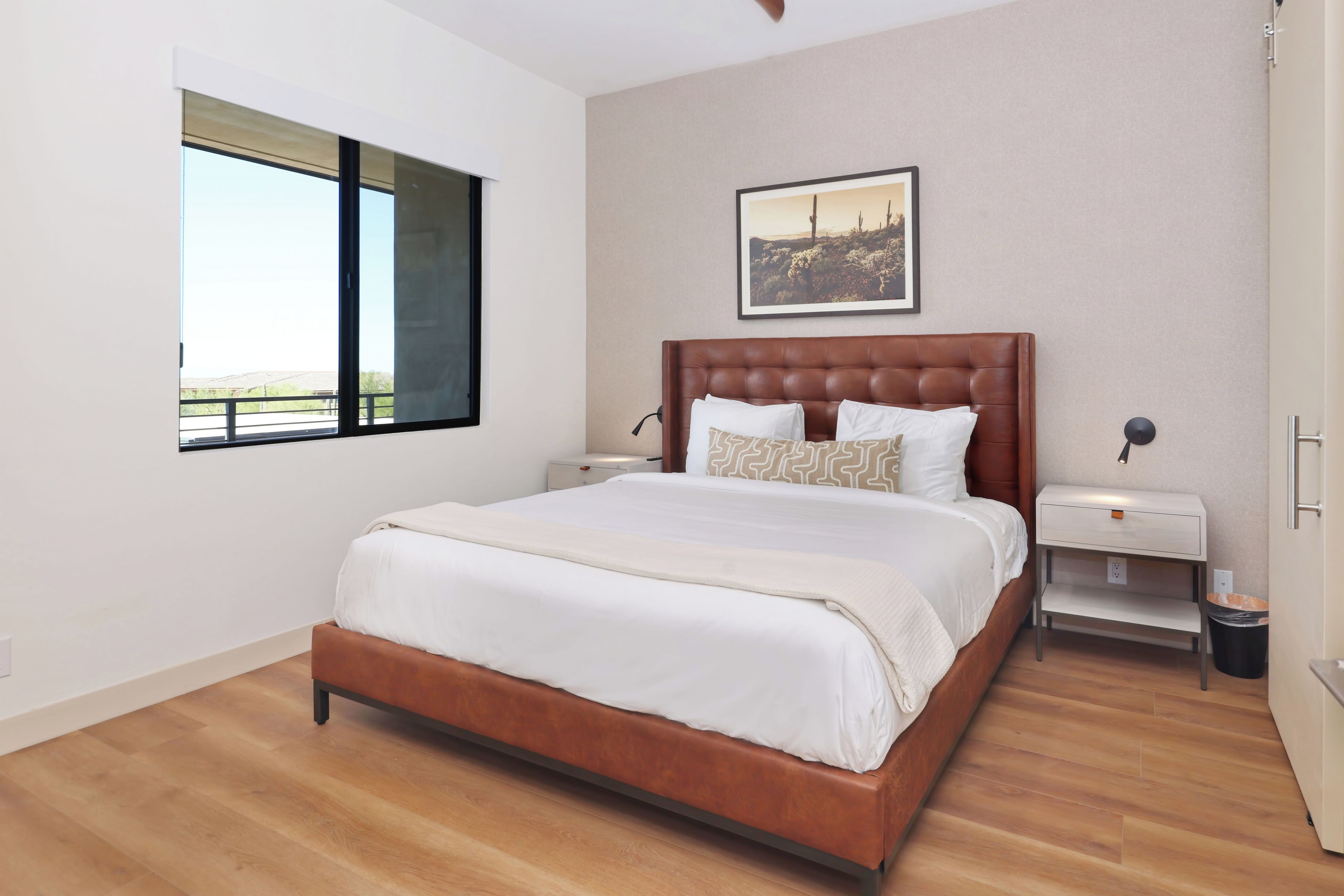 A bedroom with a large bed, a nightstand, a wall-mounted lamp, a window with a view, and a framed picture on the wall.
