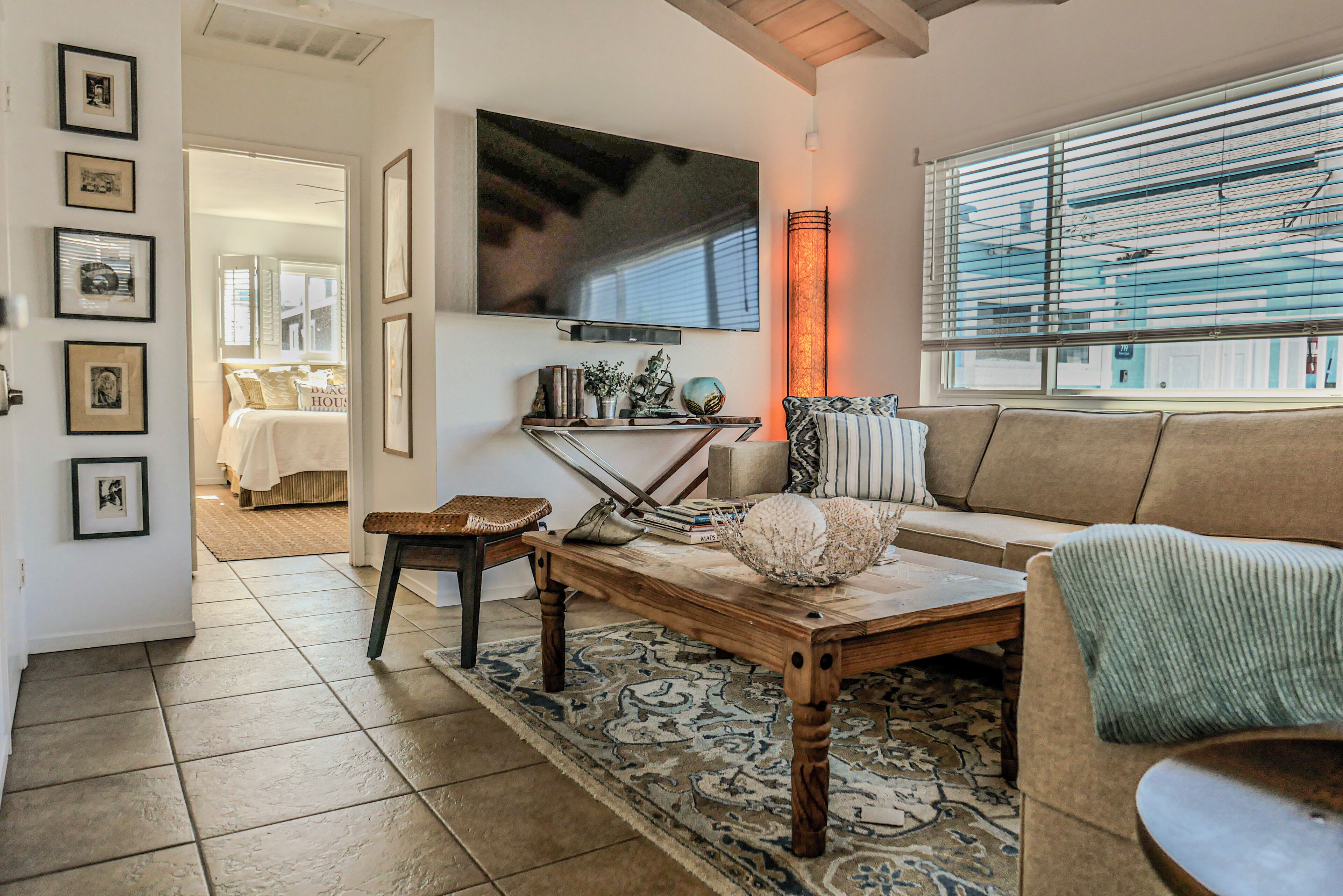 A living room with a wooden coffee table, a sofa, a television mounted on the wall, and a large window with blinds.