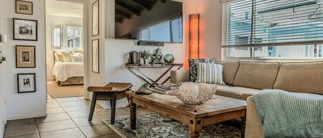 A living room with a wooden coffee table, a sofa, a television mounted on the wall, and a large window with blinds.