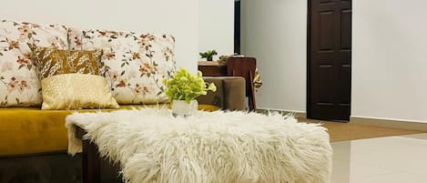 A modern living room with a sofa, a fluffy white pouf, a ceiling fan, and a dark wooden door.