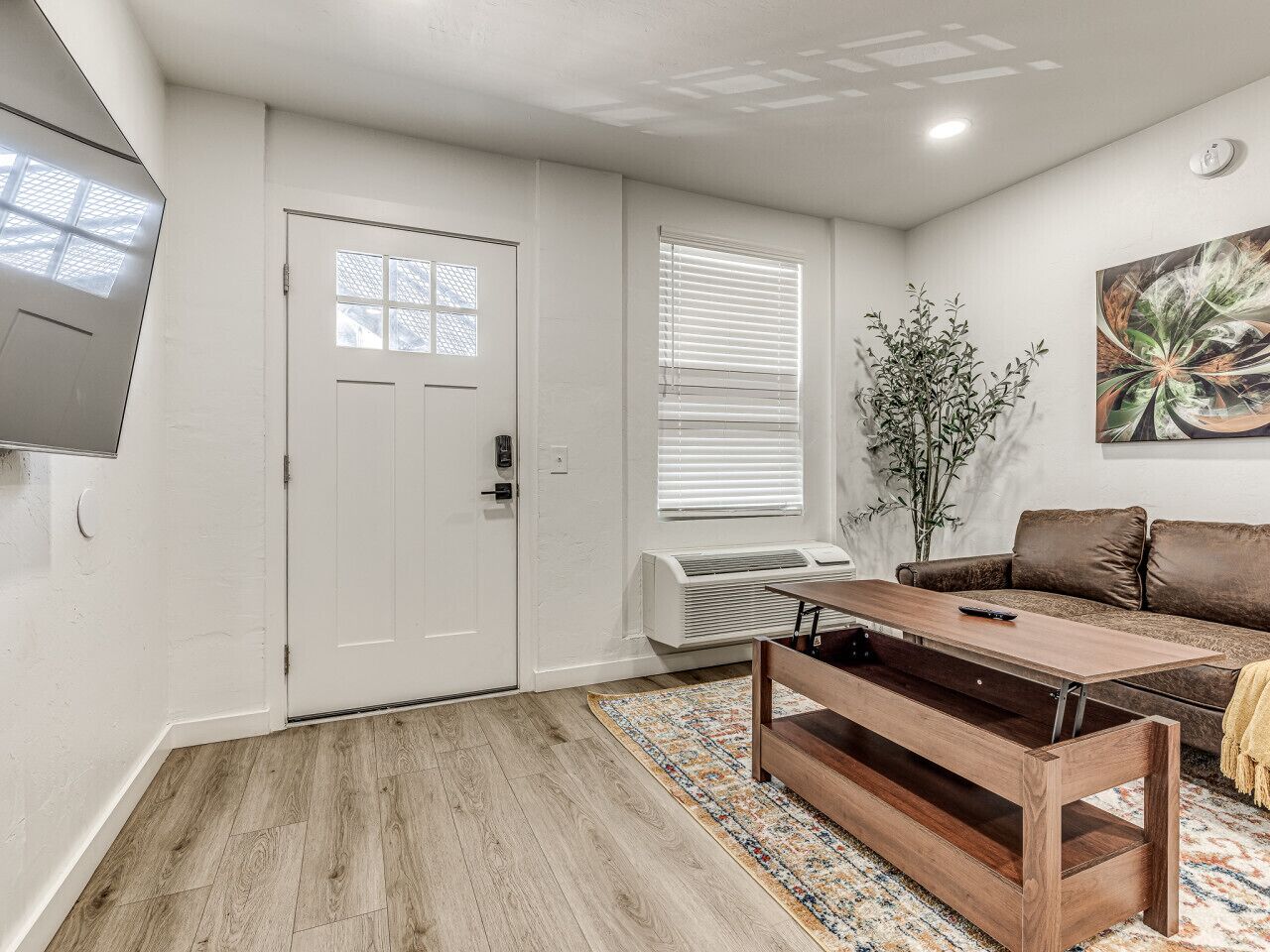 A living room with a wooden floor, a brown sofa, a coffee table, a TV mounted on the wall, and a door with a window.
