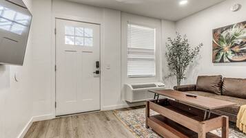 A living room with a wooden floor, a brown sofa, a coffee table, a TV mounted on the wall, and a door with a window.