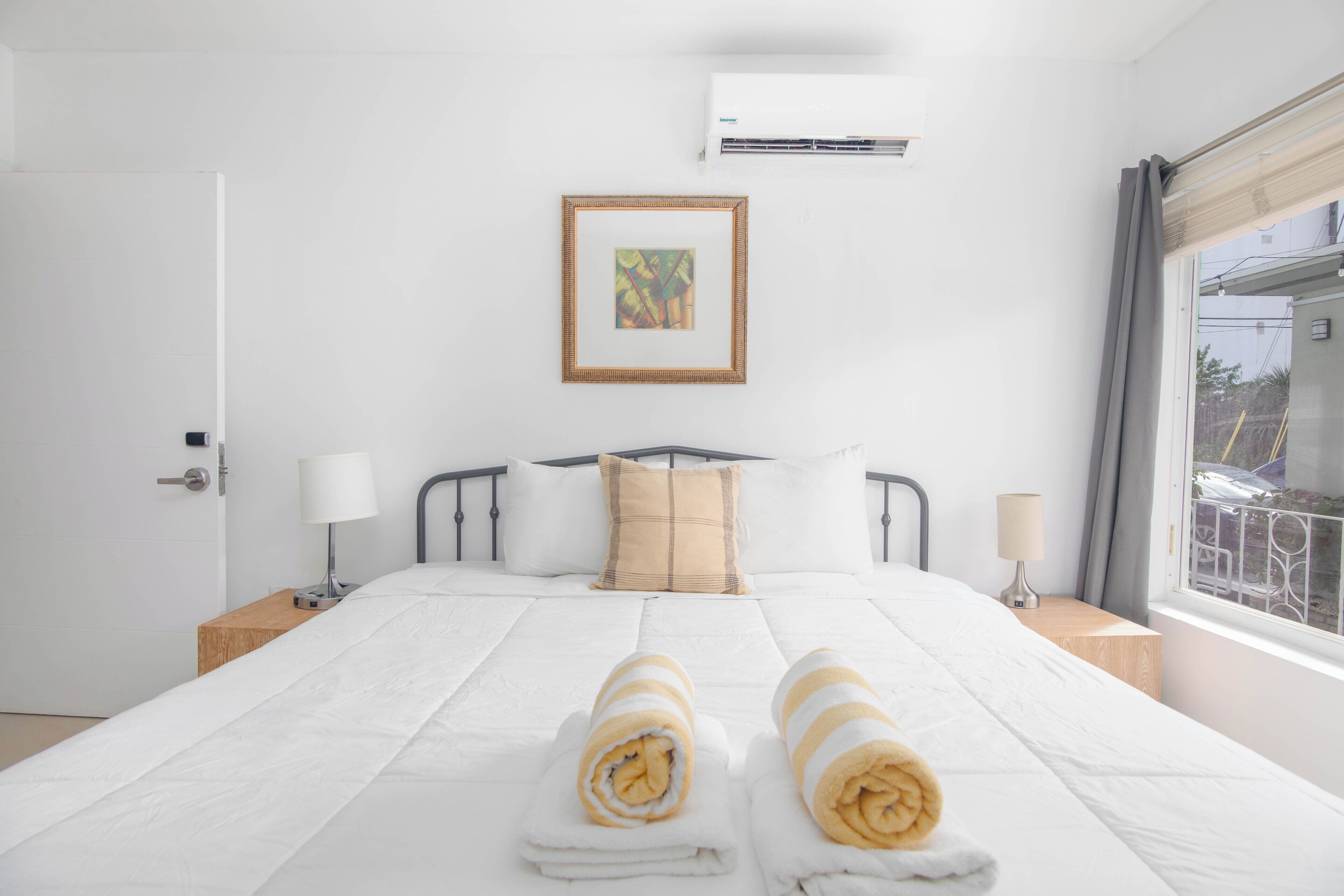 A neatly made bed with two rolled towels, a framed picture on the wall, and a window with curtains.