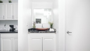 A modern kitchen with white cabinets, a black countertop, a copper sink, and a mirror above it. - Boutique Apartments Flamingo (Miami Beach)