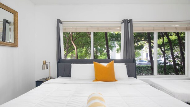 A hotel room with a large bed, a smaller bed, a window with curtains, a nightstand with a lamp, and a mirror on the wall. - Boutique Apartments Flamingo (Miami Beach)