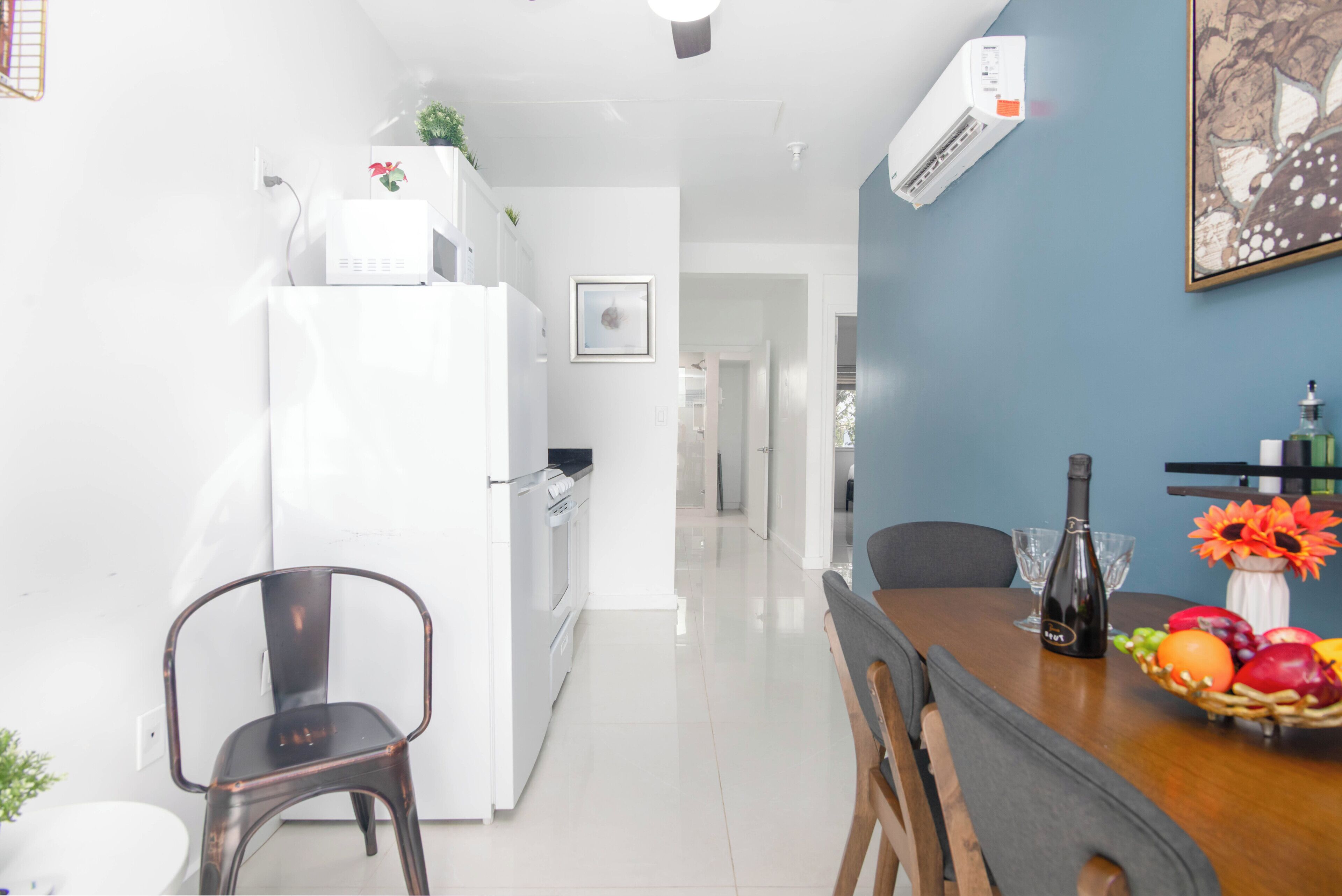 Una cocina moderna con electrodomésticos blancos, una zona de comedor con mesa y sillas de madera, y una pared azul decorativa.