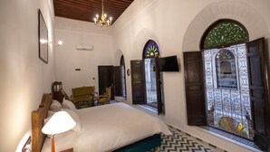 A bedroom with a bed, a lamp, a chair, and a window with decorative ironwork. - Riad SOULTANA every stay tells a story (Fes)