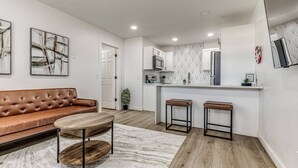 A modern kitchen with white cabinets, a microwave, and a sink. There is a brown leather sofa and two stools in the living area. - Remodeled Modern 1 Bed (Oklahoma City)