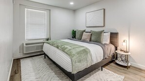 A bedroom with a bed, a bedside table with a lamp, a radiator, and a window with blinds. - Remodeled Modern 1 Bed (Oklahoma City)