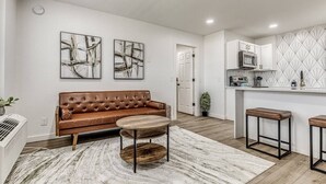 A modern living room with a brown leather sofa, a round coffee table, and two bar stools. A kitchen with white cabinets and a patterned backsplash is visible in the background. - Remodeled Modern 1 Bed (Oklahoma City)
