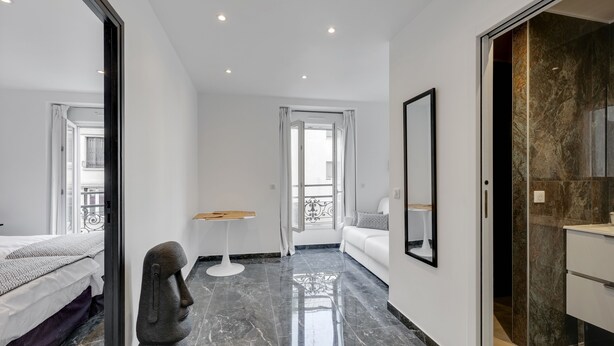 A modern hotel room with a bed, a small table, a chair, a mirror, and a bathroom.