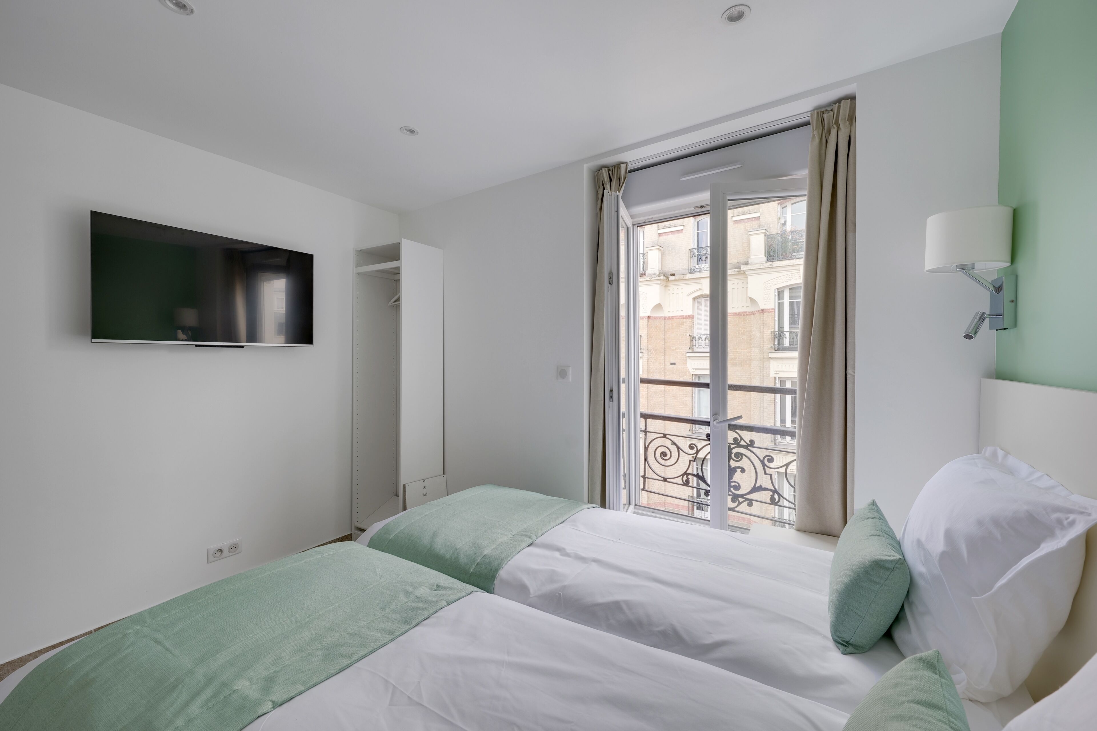 A bedroom with two beds, a TV mounted on the wall, a window with curtains, and a balcony view.