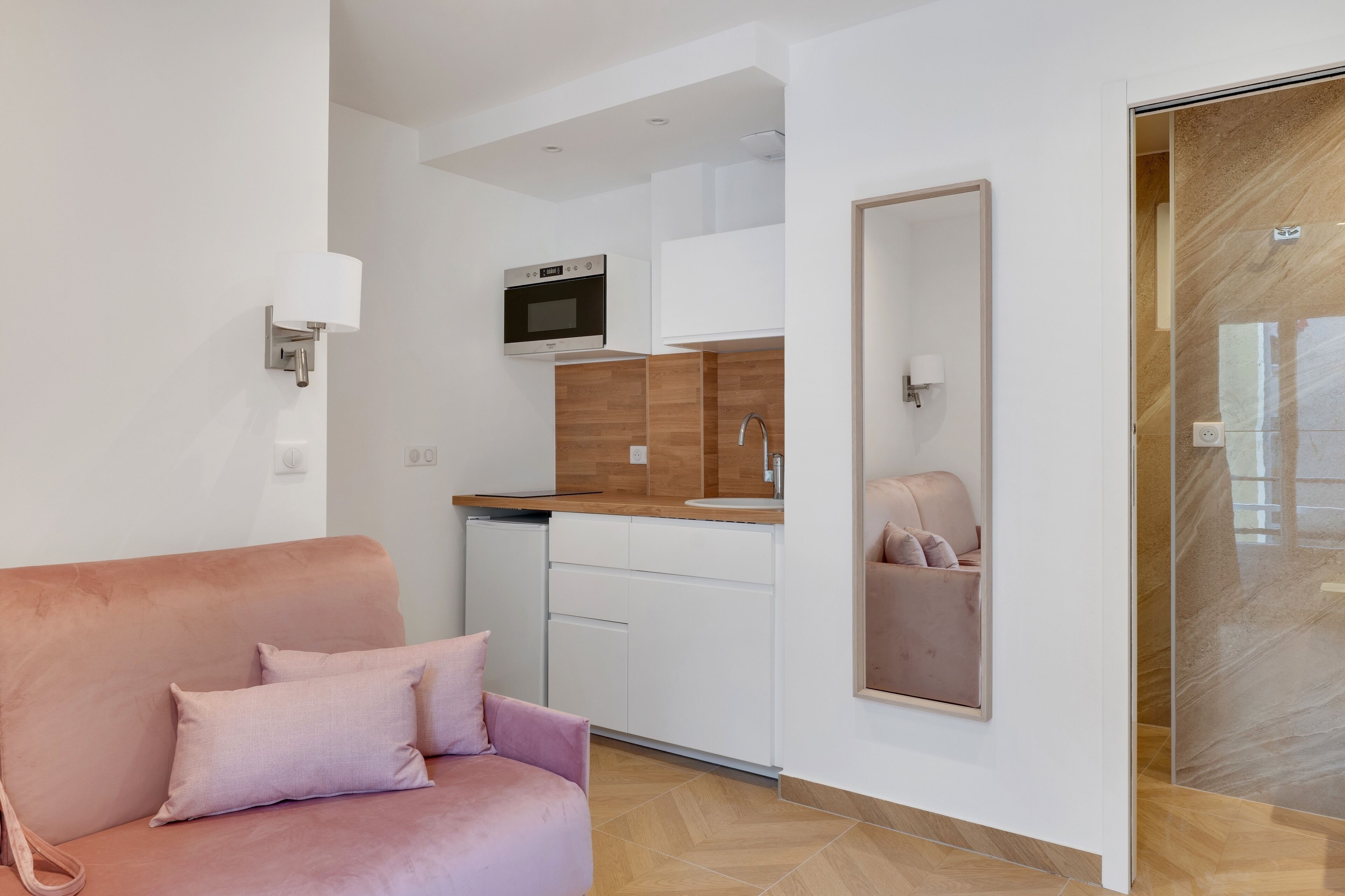 A modern kitchen with white cabinets, a microwave, and a wooden countertop. There is a pink sofa with cushions in the living area.
