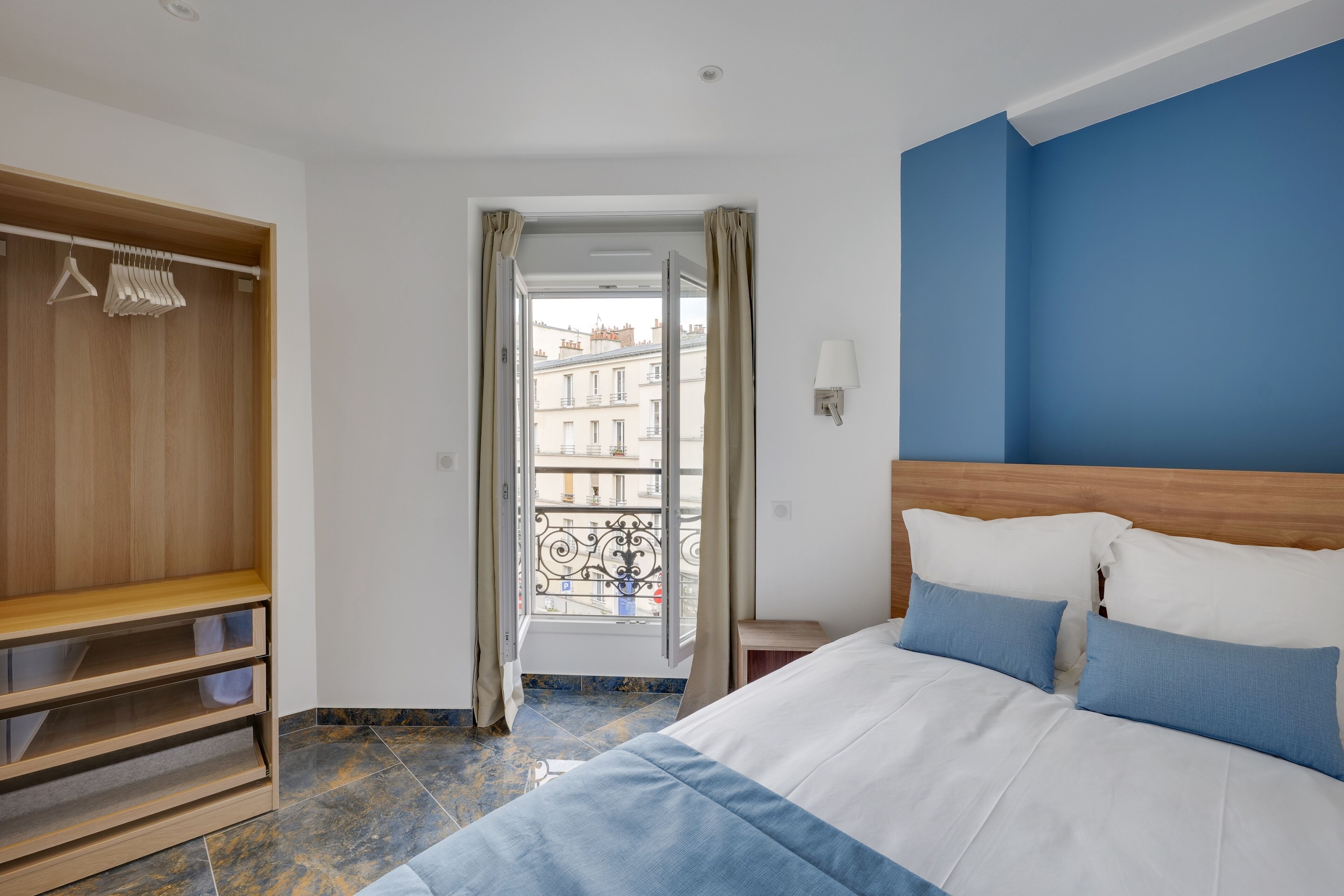 A bedroom with a bed, a wooden wardrobe, and a view of buildings through an open door.