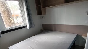 Une petite chambre avec un lit simple, une tête de lit en bois, une fenêtre avec un rideau et des étagères intégrées au-dessus du lit.