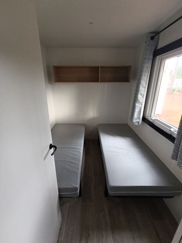A small, narrow room with two single beds aligned against the wall, a window with patterned curtains, and a wooden shelf above the beds.