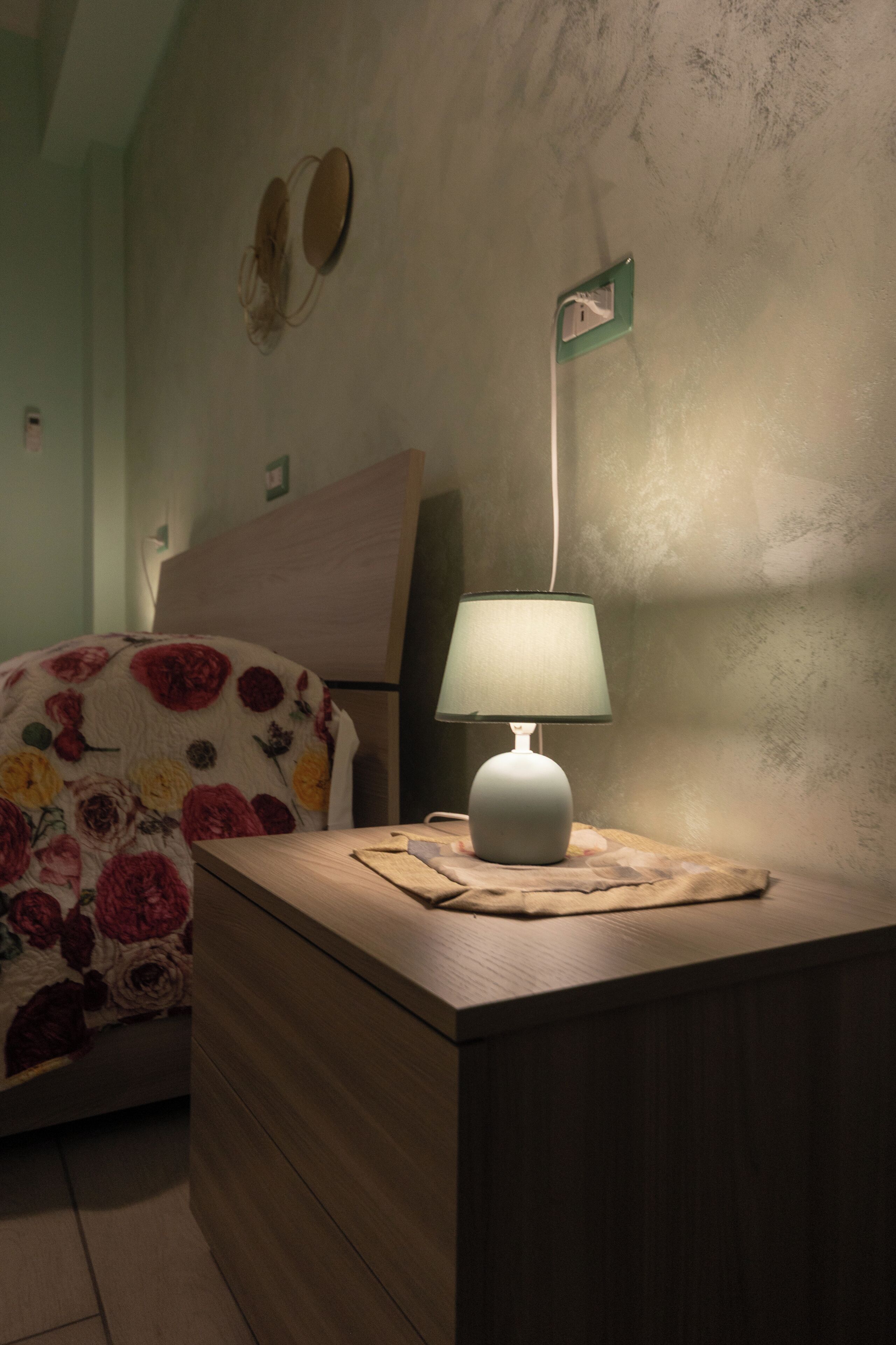 A bedroom with a bed covered in a floral blanket, a bedside table with a lamp, and a wall-mounted light switch.