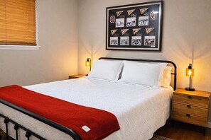 A neatly made bed with a white comforter and a red blanket, a framed collage of sports memorabilia on the wall, and two bedside lamps on wooden nightstands.